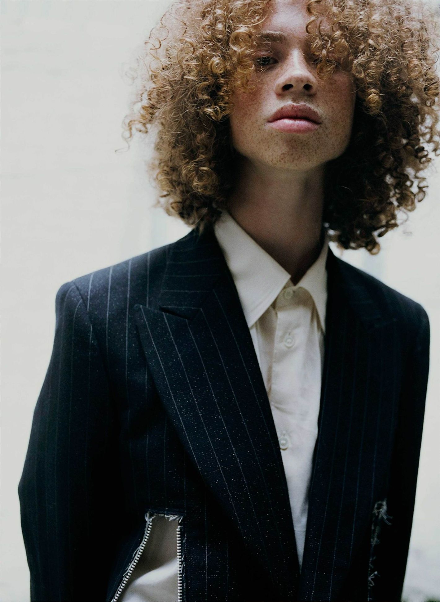 Portrait of a light-skinned adult male with curly hair wearing a pinstripe blazer and white button-down shirt against a neutral background.
