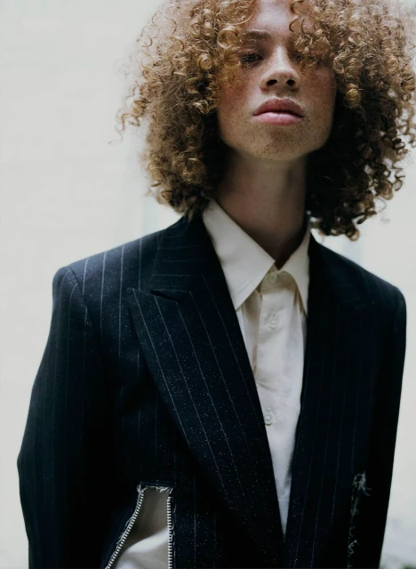 Portrait of a light-skinned adult male with curly hair wearing a pinstripe blazer and white button-down shirt against a neutral background.