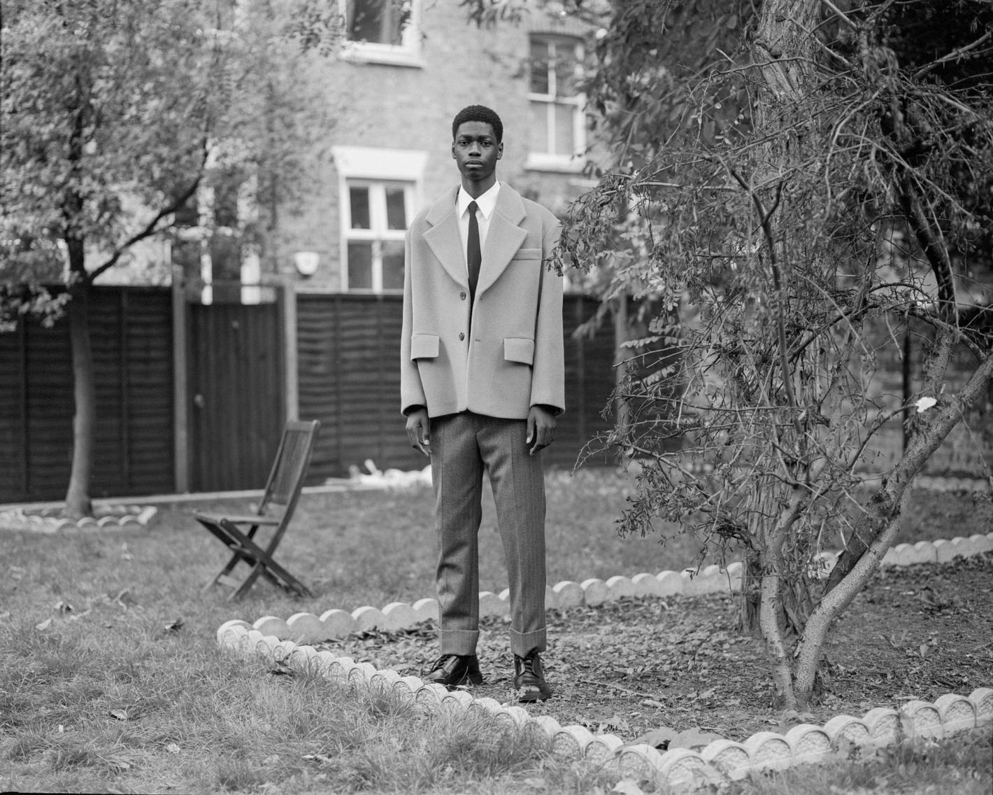 Black male model in oversized tan jacket and gray trousers standing in garden with chair, fence, and house in the background.