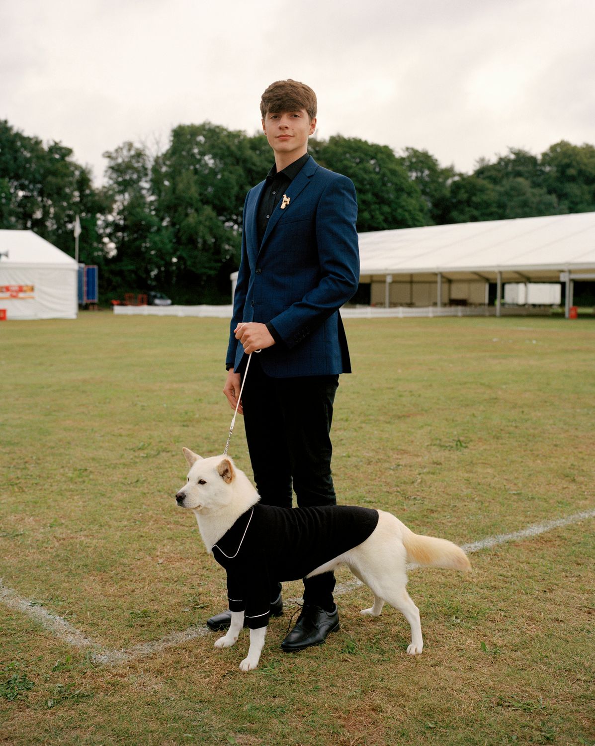 Young man in a suit stands with a white dog in a black sweater on a grassy field, with tents in the background on an overcast day.
