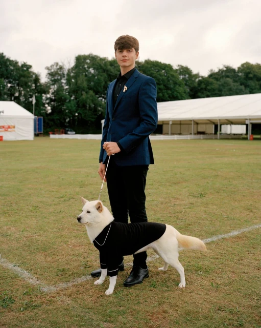 Young man in a suit stands with a white dog in a black sweater on a grassy field, with tents in the background on an overcast day.