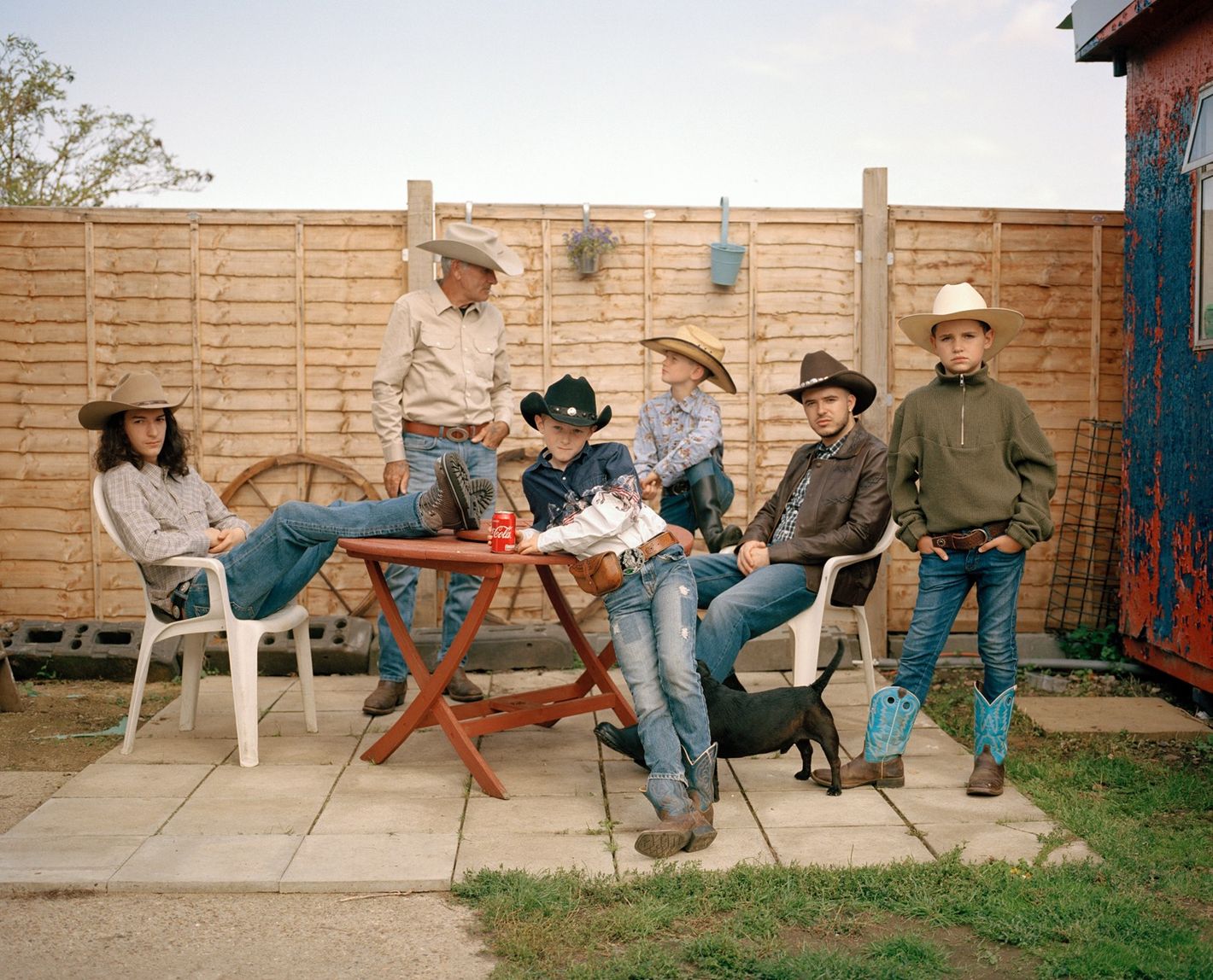 Outdoor portrait of six people in cowboy attire and a dog posing on a patio, featuring a wooden fence and a glimpse of a red building.