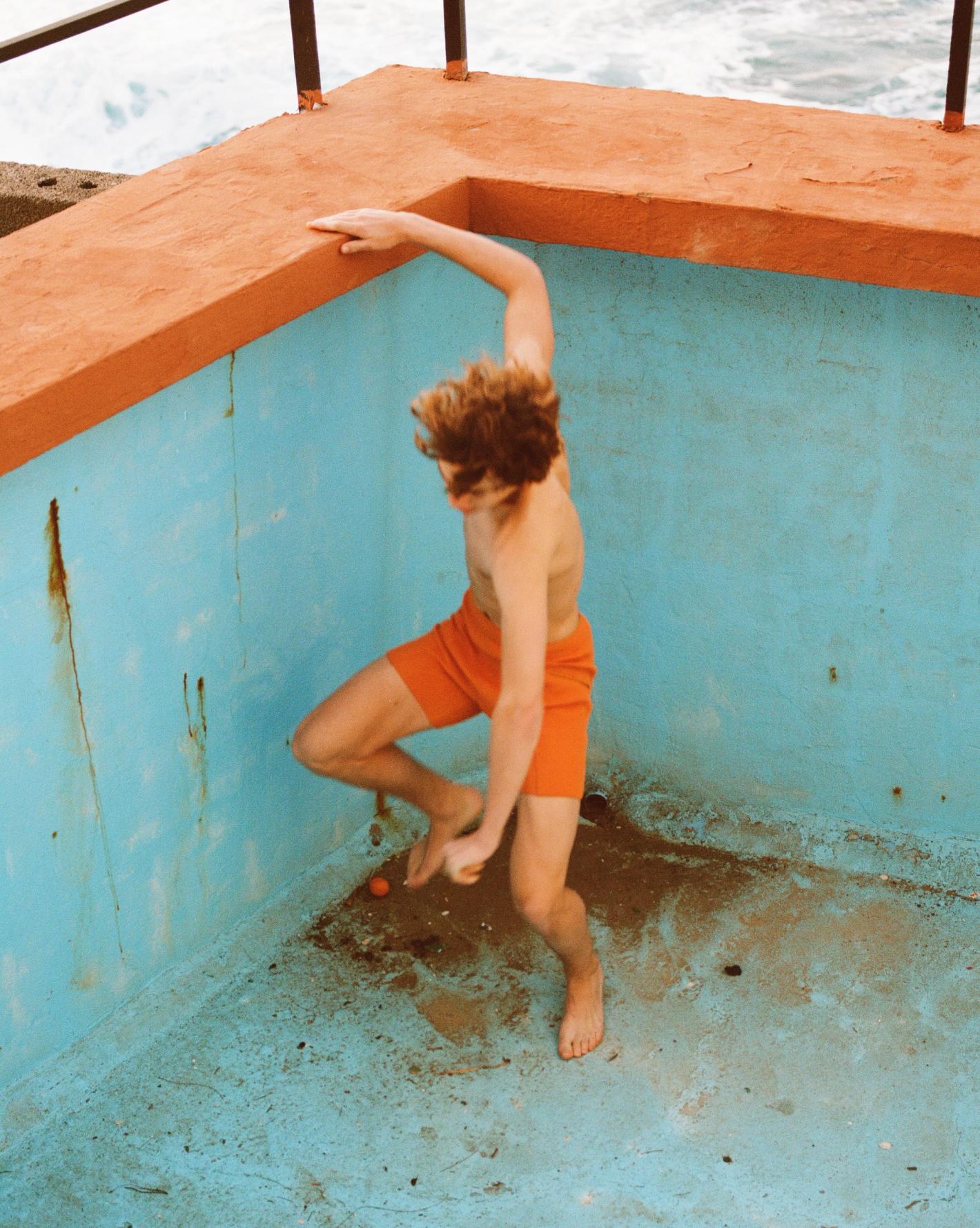 High-angle view of a young adult male with auburn hair emerging from a faded turquoise and orange swimming pool on a sunny day.
