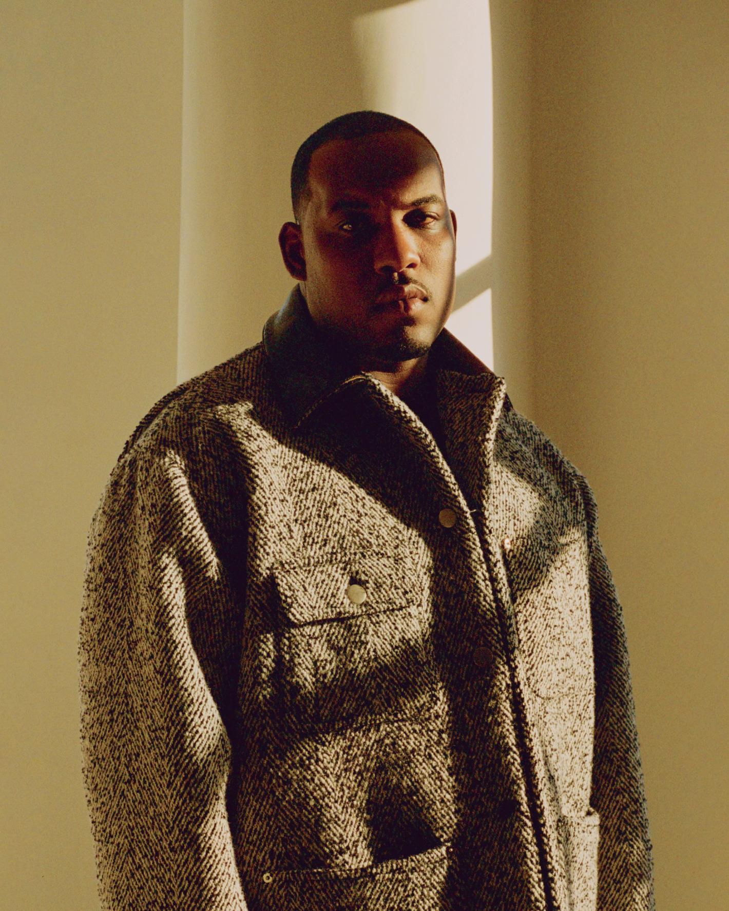 Portrait of an adult male with short dark hair wearing a tweed jacket standing indoors in a location with natural light and shadows.