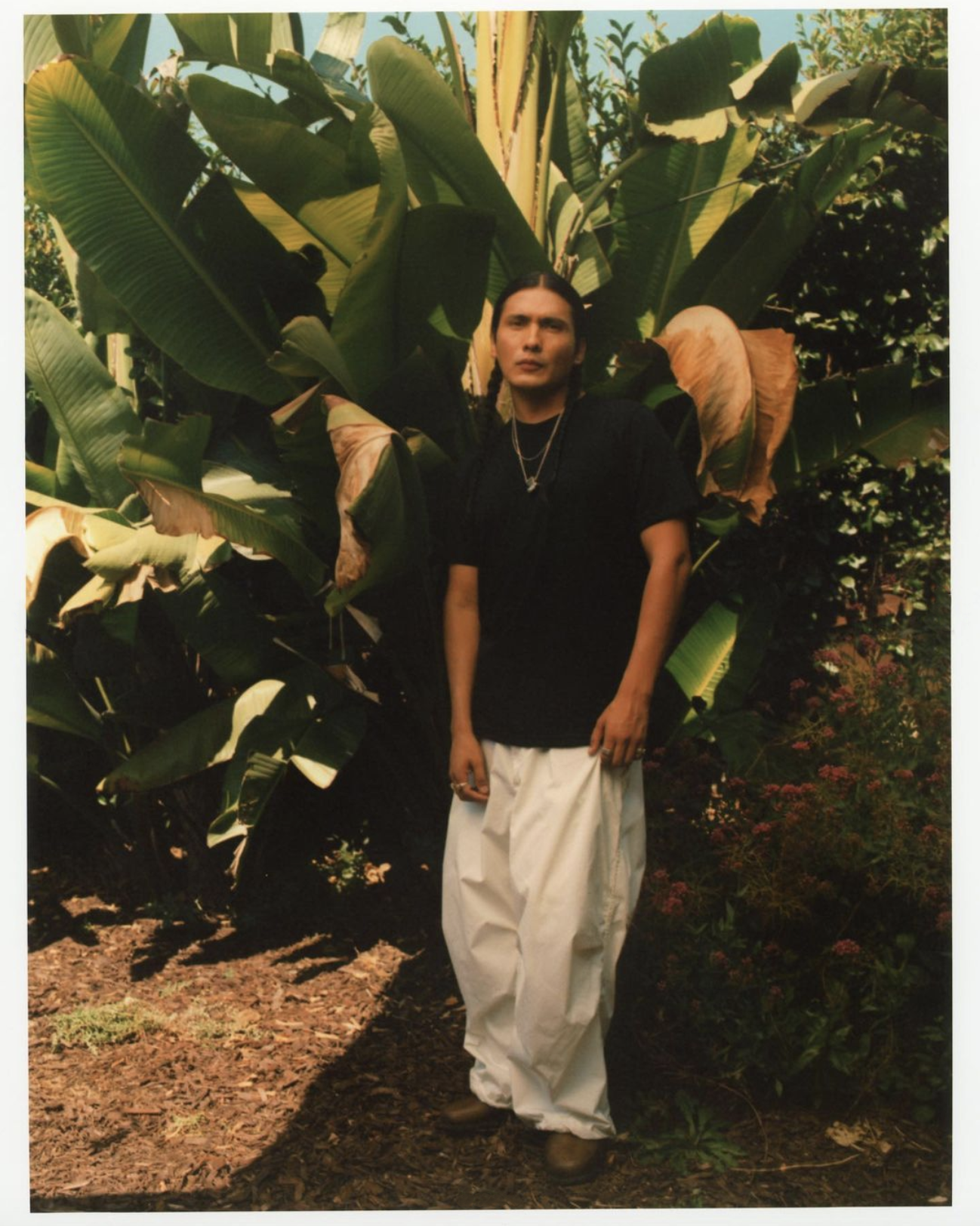 A stylish person stands in front of large green leafy plants, wearing a black t-shirt and white pants in an outdoor setting.