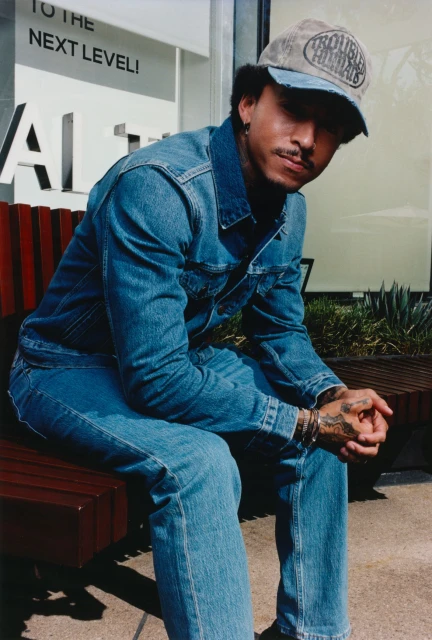 A man with tattoos wearing a denim jacket and cap, sits on a bench in an urban environment with muted natural light, showcasing casual fashion.