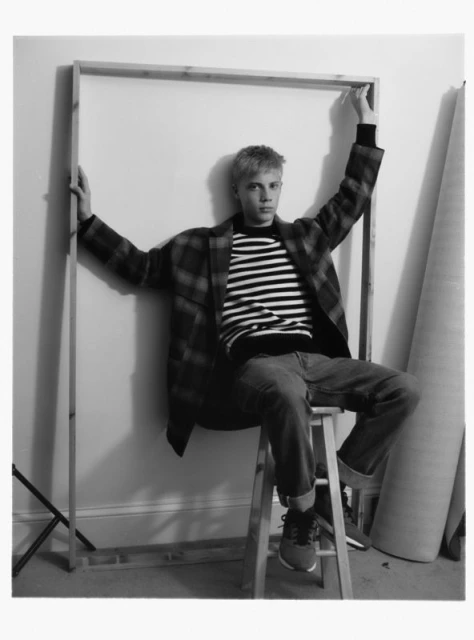 Blonde male model seated on stool, holding up wooden frame, wearing striped shirt, blue jeans, and plaid coat indoors.