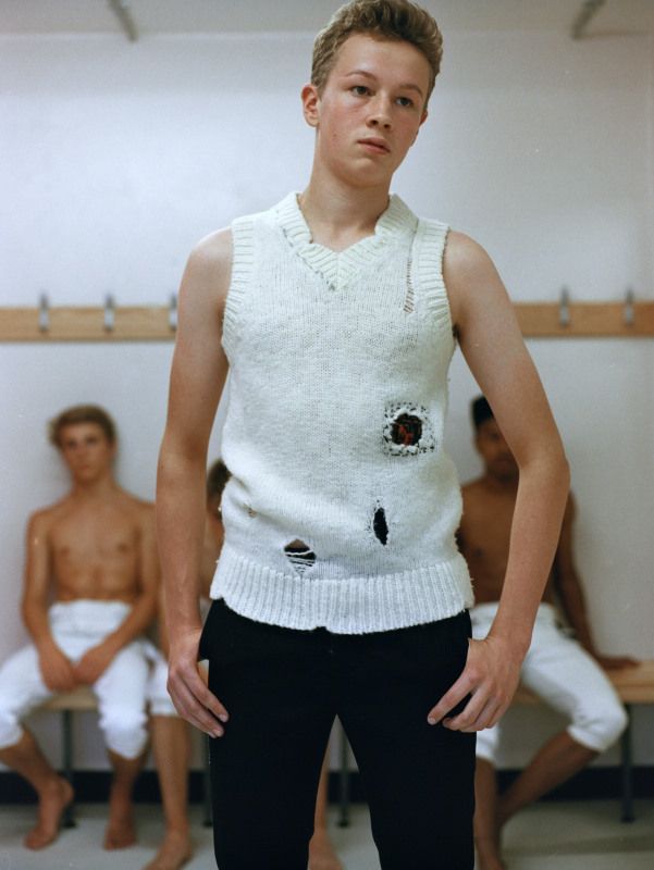 Blonde male wearing a ripped white knitted vest with black trousers, standing in front of two shirtless men on a bench.