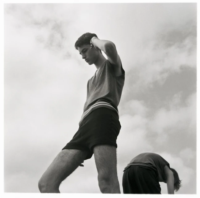 Two young adult males pose outside against a cloudy sky, one standing, one bent over, in a high-contrast black-and-white outdoor photograph.
