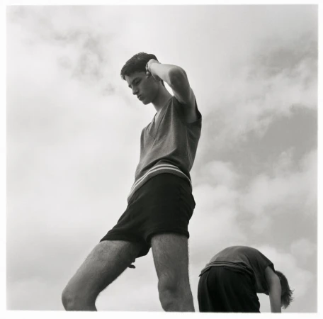 Two young adult males pose outside against a cloudy sky, one standing, one bent over, in a high-contrast black-and-white outdoor photograph.
