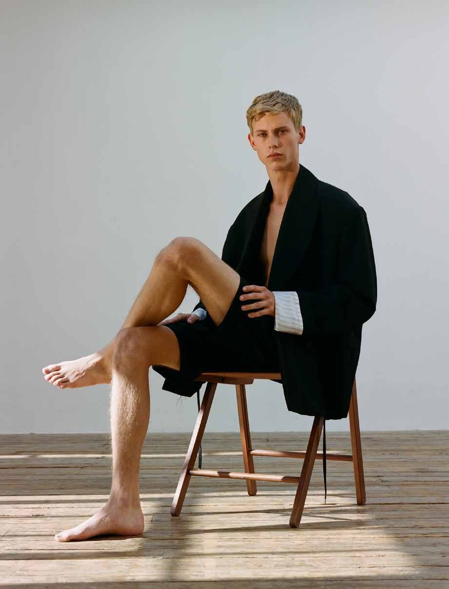 Blond male model sitting on wooden chair in photo studio wearing black blazer, shorts, and bare feet, against white backdrop and wood floor.