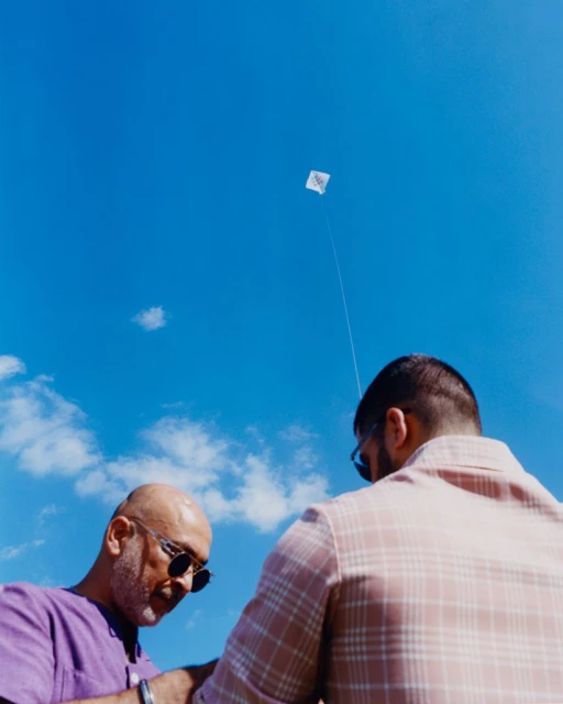 Two adult men flying a white kite in a clear blue sky with scattered clouds, viewed from a low angle in bright daylight.