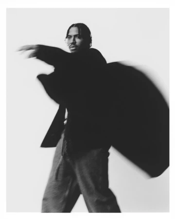 Monochrome fashion portrait of a man in motion wearing a dark jacket and trousers against a white seamless backdrop.