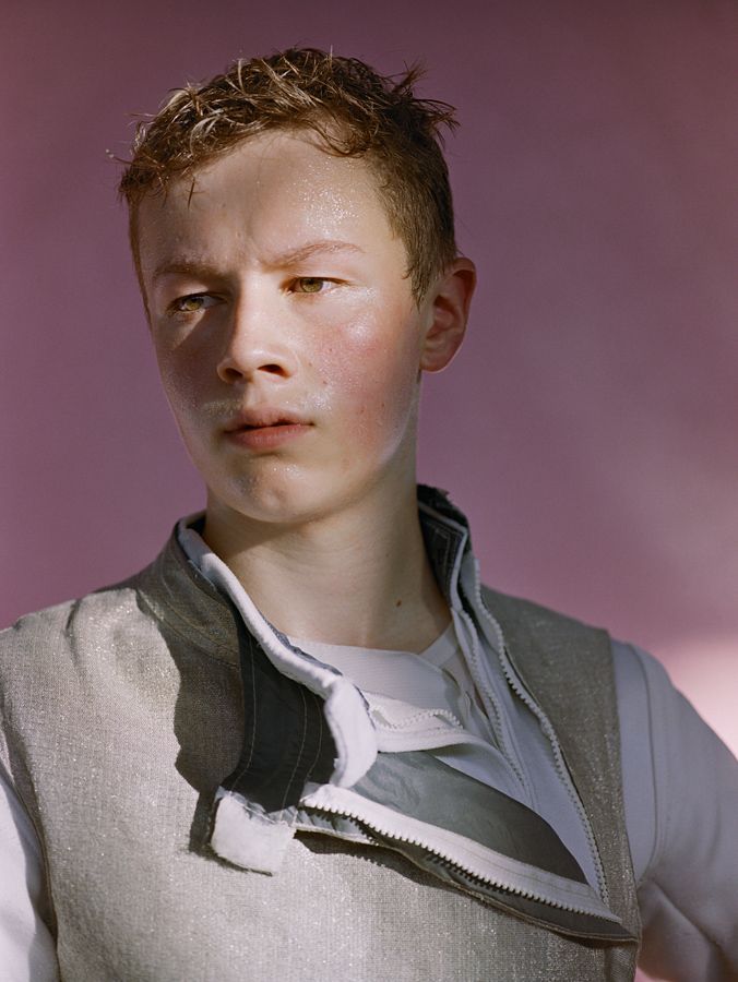 Portrait of a young adult in a light grey vest with a partly unzipped zipper, with a pink background and soft, diffused lighting.