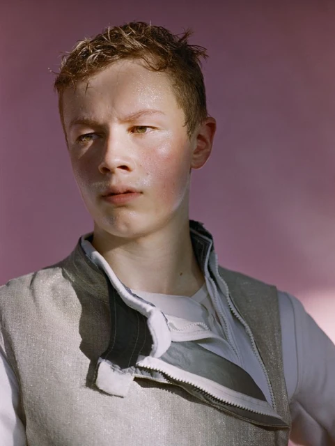 Portrait of a young adult in a light grey vest with a partly unzipped zipper, with a pink background and soft, diffused lighting.