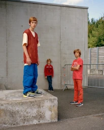 Three boys posing outdoors against a concrete wall, styled by Adam Winder with a red knit vest and blue trousers.