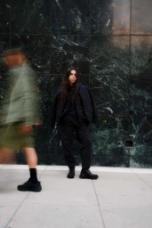 Woman in dark clothing posing against a green marble wall with a blurred figure walking past in the foreground.