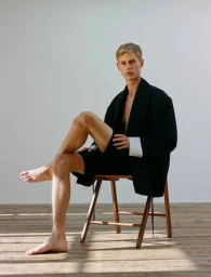 Blonde male model seated on a wooden chair wearing a black coat and shorts against a white wall.