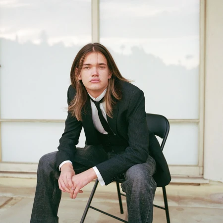 Young man with long hair wearing a black jacket and white shirt seated on a chair