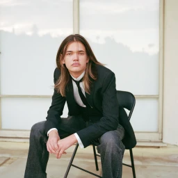 Young man with long hair wearing a black jacket and white shirt seated on a chair