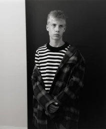 Young man with short blonde hair wearing a striped crewneck sweater and plaid jacket against a split backdrop