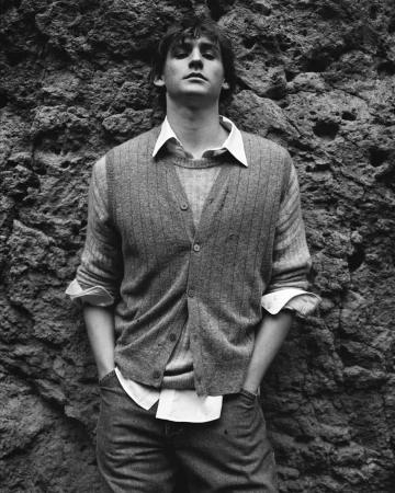 Monochrome portrait of a young man wearing a knit vest and white shirt against a textured stone wall