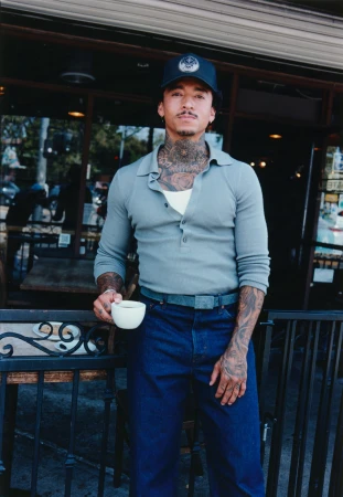 Man with neck tattoos wearing a grey polo shirt holding a white coffee cup outside a storefront.