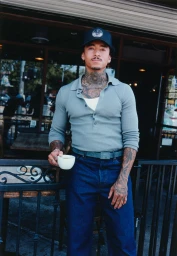 Man with neck tattoos wearing a grey polo shirt holding a white coffee cup outside a storefront.