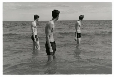 Three young men wading in shallow ocean water under an overcast sky, styled by Adam Winder.