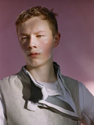 Close-up portrait of a young man with damp hair wearing a silver fencing vest against a pink background