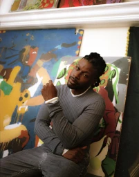 Black man with braided hair wearing a grey ribbed sweater seated against large colourful paintings