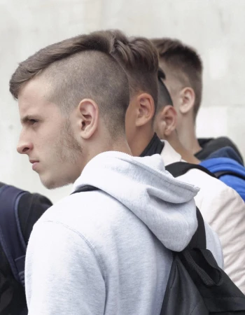 Profile portrait of a young man wearing a light grey hoodie and a black backpack, with other young men lined up behind him.