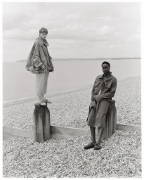 Two male models posing on wooden posts along a pebbled beach under an overcast sky, styled by Adam Winder.