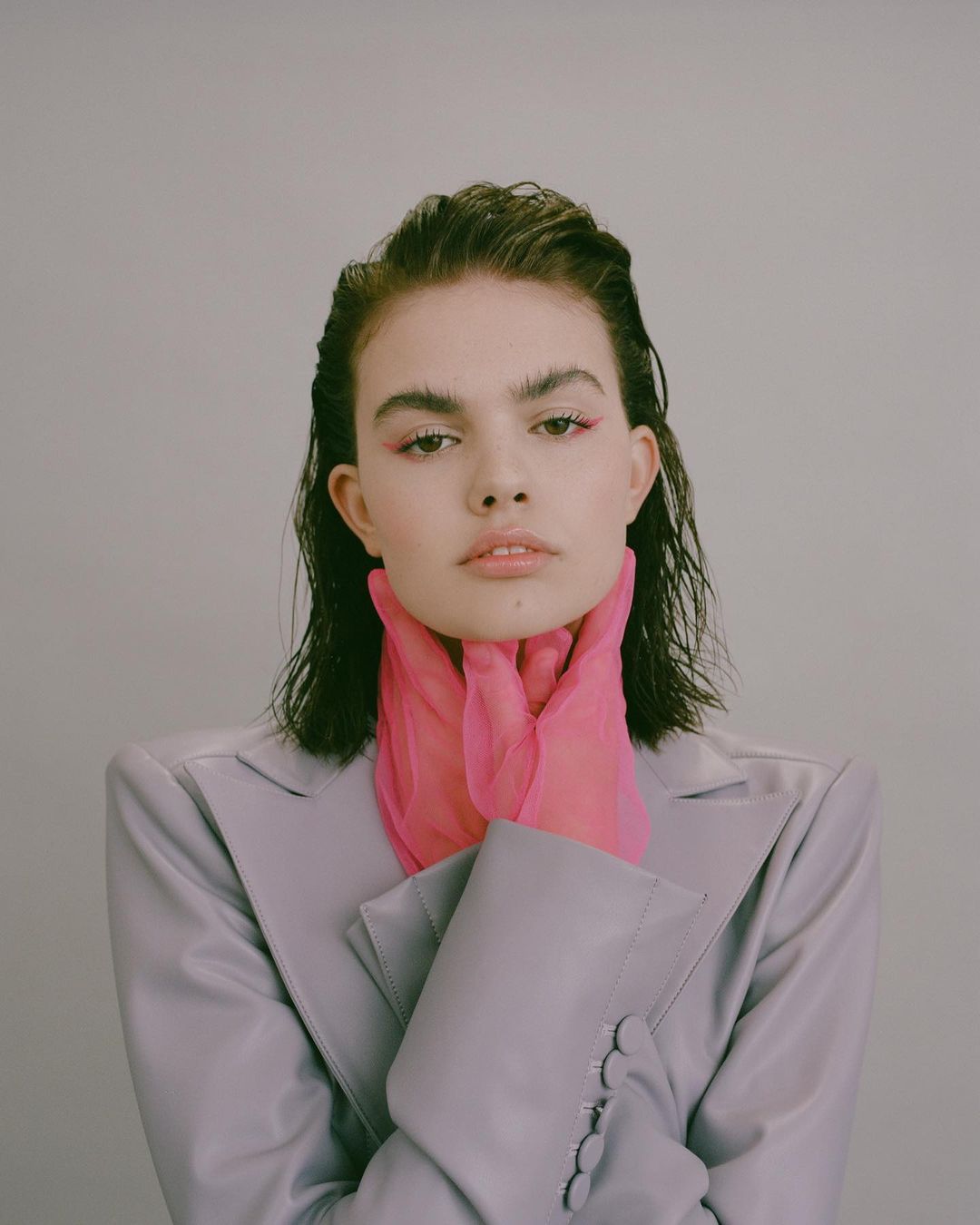 Portrait of a young woman with pink eye shadow and pink transparent gloves, wearing a grey blazer against a pale grey background.