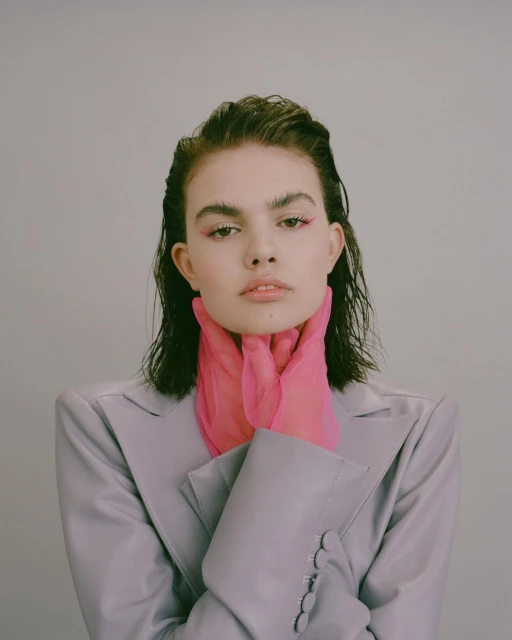 Portrait of a young woman with pink eye shadow and pink transparent gloves, wearing a grey blazer against a pale grey background.