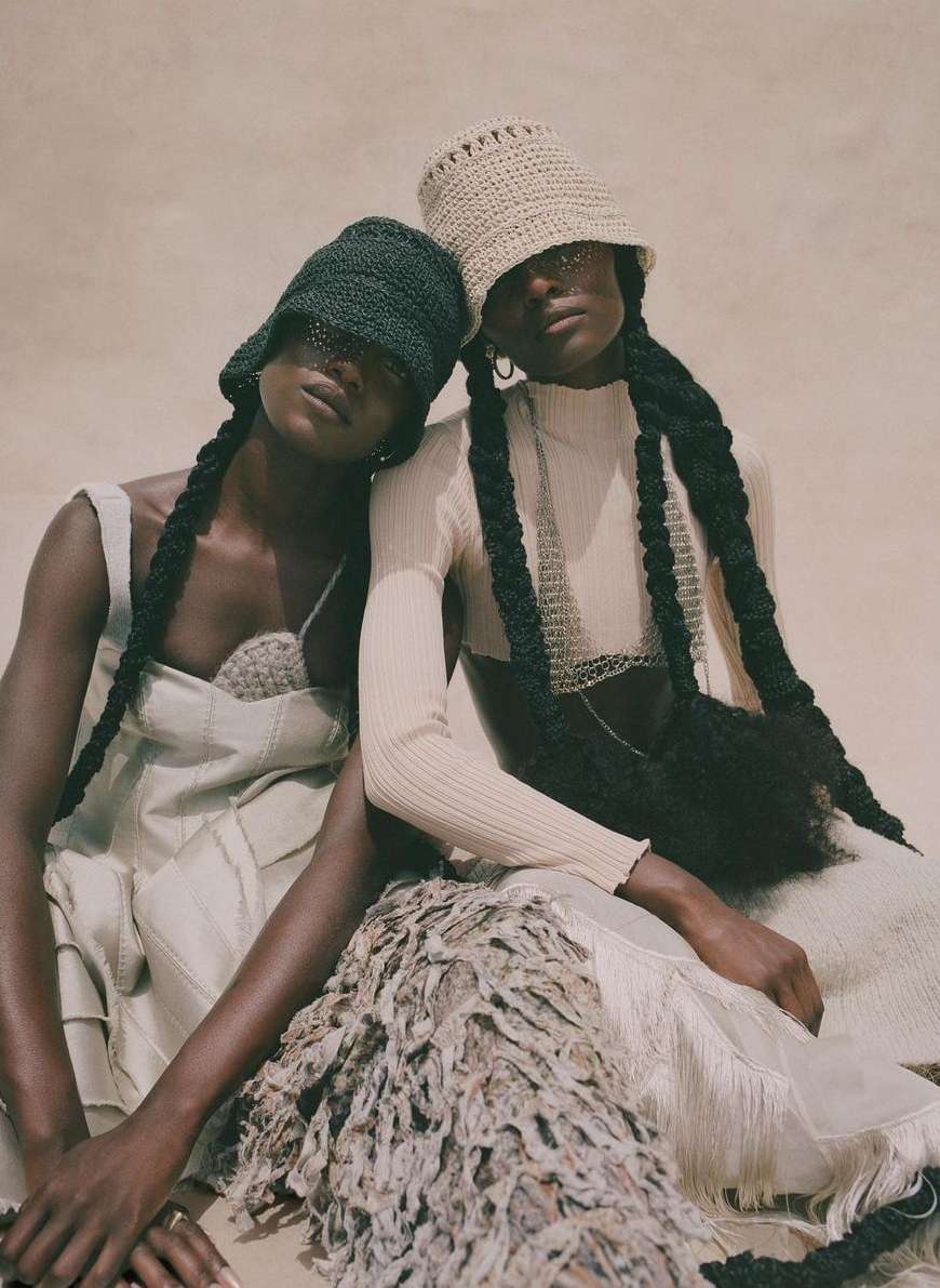 Two women pose in matching neutral outfits and crocheted hats, showcasing unique braided hairstyles, indoor setting with soft, diffused light.