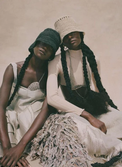 Two women pose in matching neutral outfits and crocheted hats, showcasing unique braided hairstyles, indoor setting with soft, diffused light.