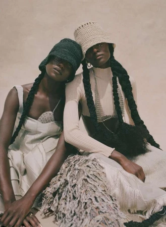 Two women pose in matching neutral outfits and crocheted hats, showcasing unique braided hairstyles, indoor setting with soft, diffused light.