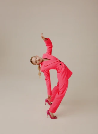 Model in a hot pink pantsuit bending backward with one arm raised, wearing pink stiletto heels against a plain neutral background.