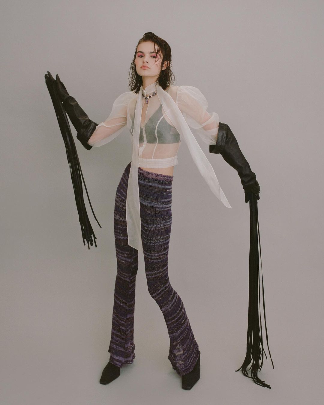 A stylish young woman poses in a sheer top, knit pants, and gloves with long fringe, standing against a pale grey studio backdrop.