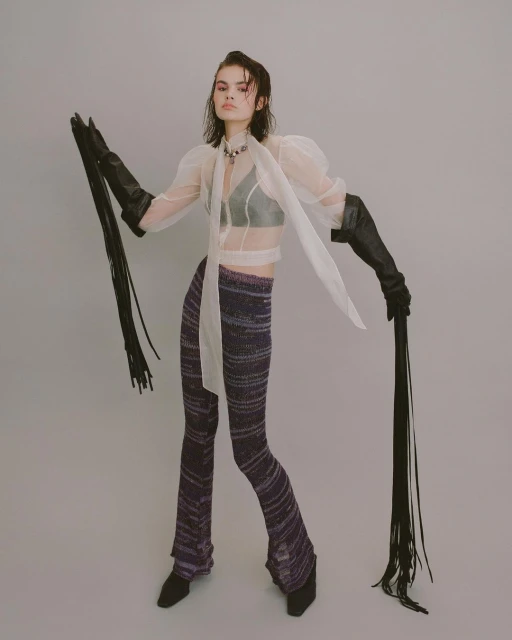 A stylish young woman poses in a sheer top, knit pants, and gloves with long fringe, standing against a pale grey studio backdrop.