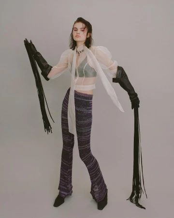 A stylish young woman poses in a sheer top, knit pants, and gloves with long fringe, standing against a pale grey studio backdrop.