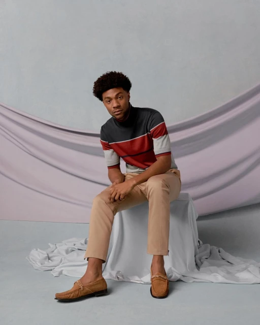 Portrait of stylish African American man in a color block top and tan pants, sitting on a white block in a studio setting.