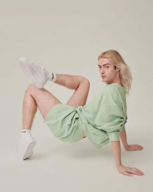 An adult posing indoors wearing a light green sweatshirt, matching shorts, white sneakers, and white socks in a studio setting.