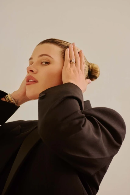 Portrait of a woman with her hands behind her head, wearing a black blazer. She has a neutral expression on her face.