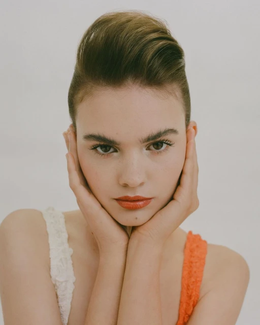 Close-up portrait of a young woman with stylized hair, facing forward, with hands gently touching her face in a brightly lit studio.