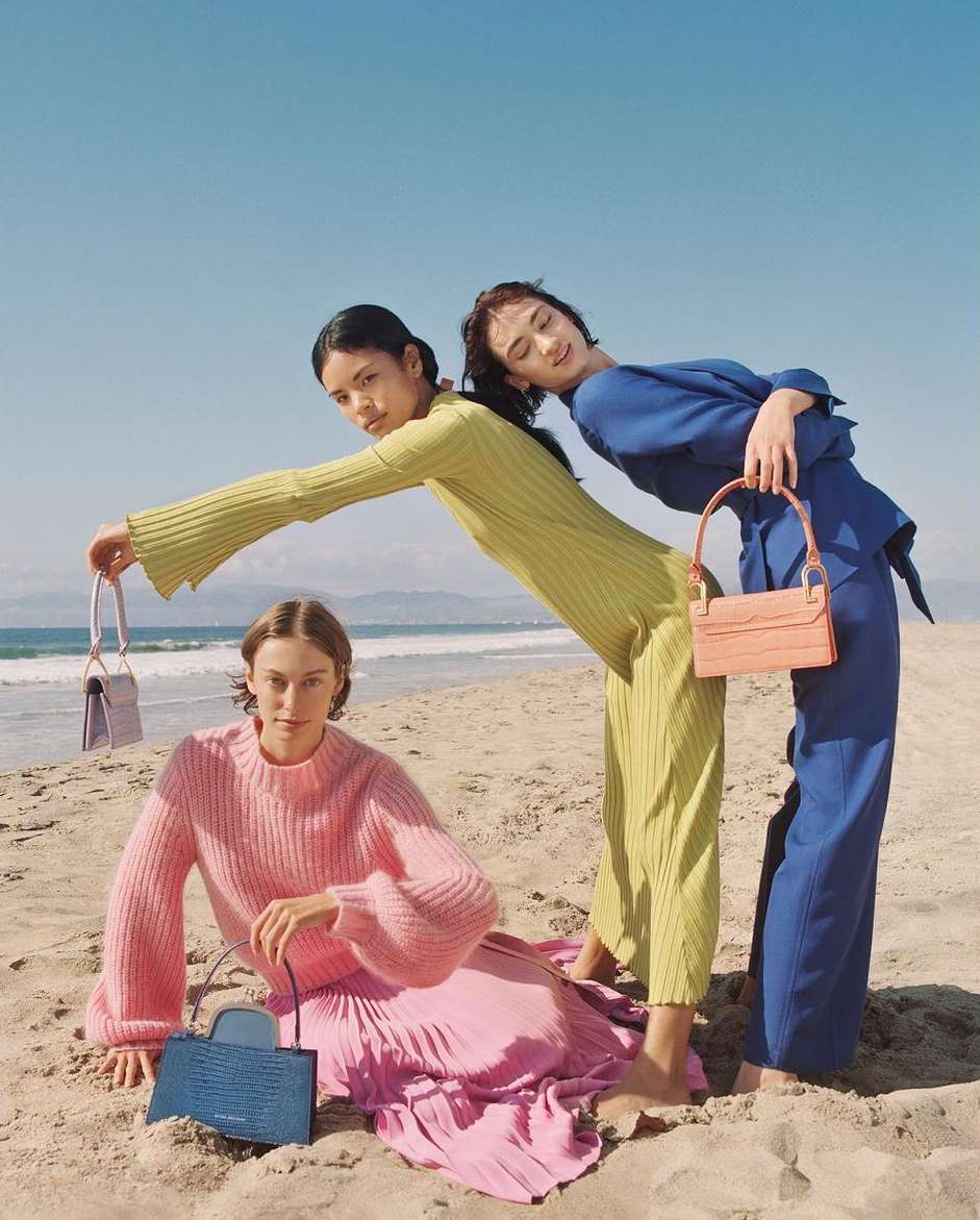 Three fashion models on a beach pose with pastel-colored handbags. The women wear coordinated knitwear in pink, blue, and green.