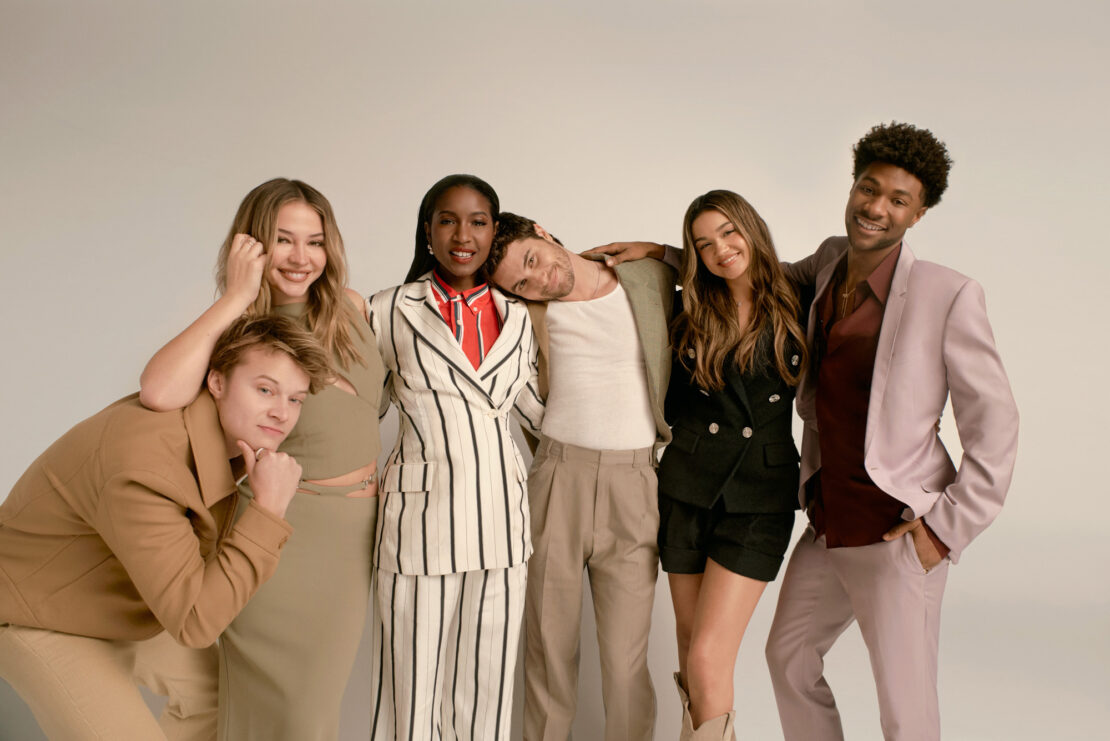 Group portrait of six young adult cast members smiling together in a bright studio. They are wearing tailored jackets and trendy casual clothing.