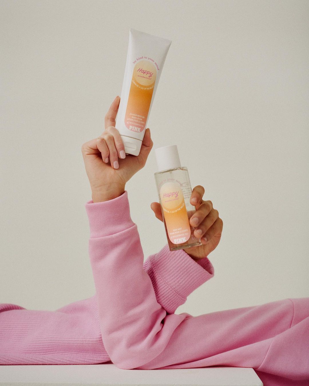 Cosmetic products are displayed and held in a studio shot featuring a long-sleeved model in pink, with lotion and spray bottles.