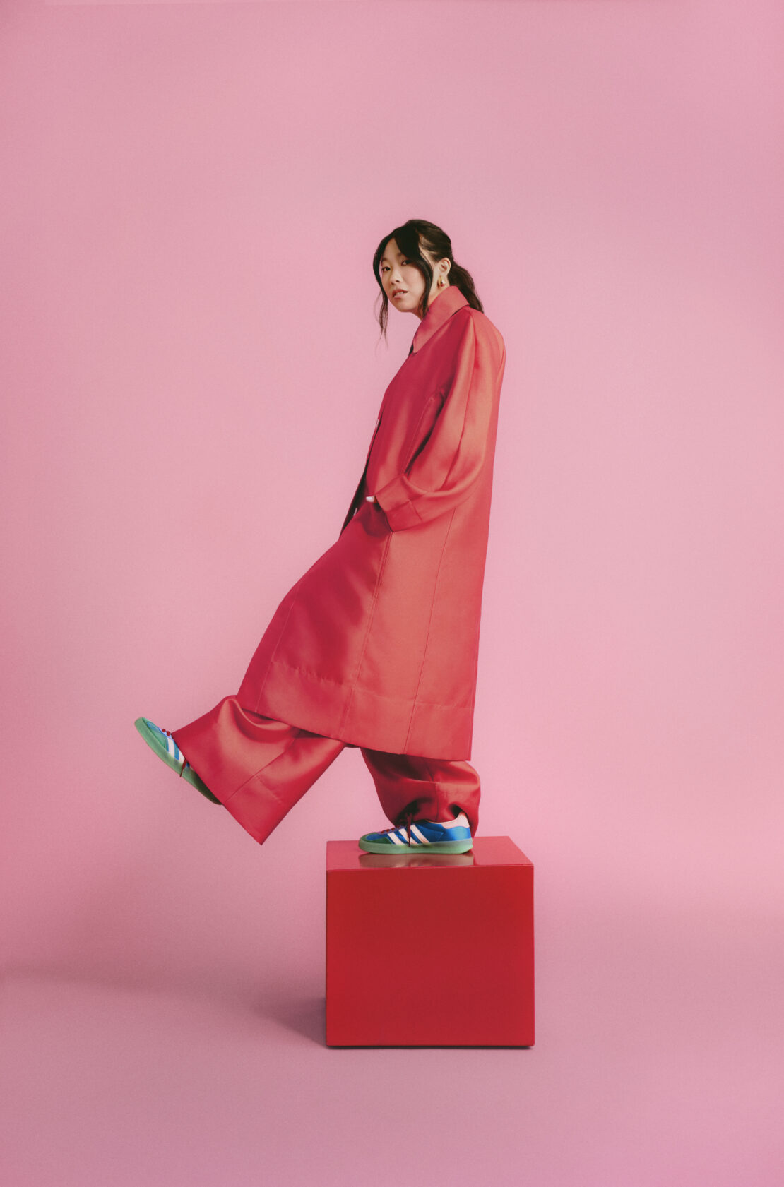 A woman in a red suit stands on a red cube against a pink backdrop, her turquoise sneaker heel lifted in dynamic pose.